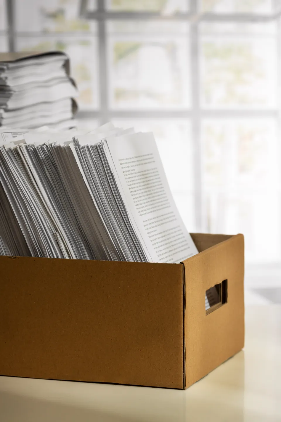 Gros plan sur une boîte en carton remplie de piles de documents papier, archives professionnelles, avec une autre pile floue en arrière-plan près d'une fenêtre.
