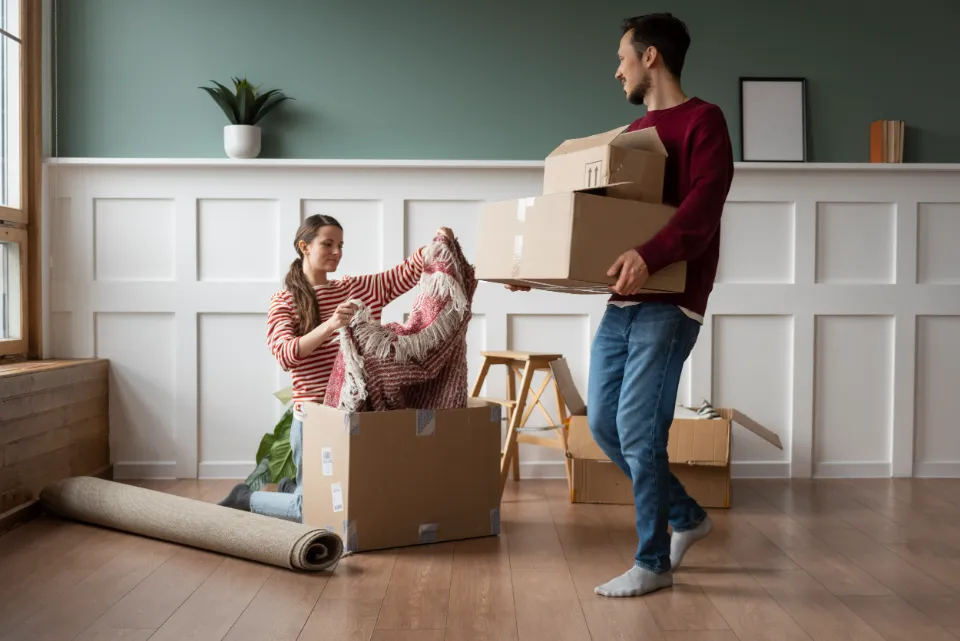 Un couple emballant des cartons pour un déménagement, l'homme porte des boîtes et la femme retire une couverture d'une boîte.