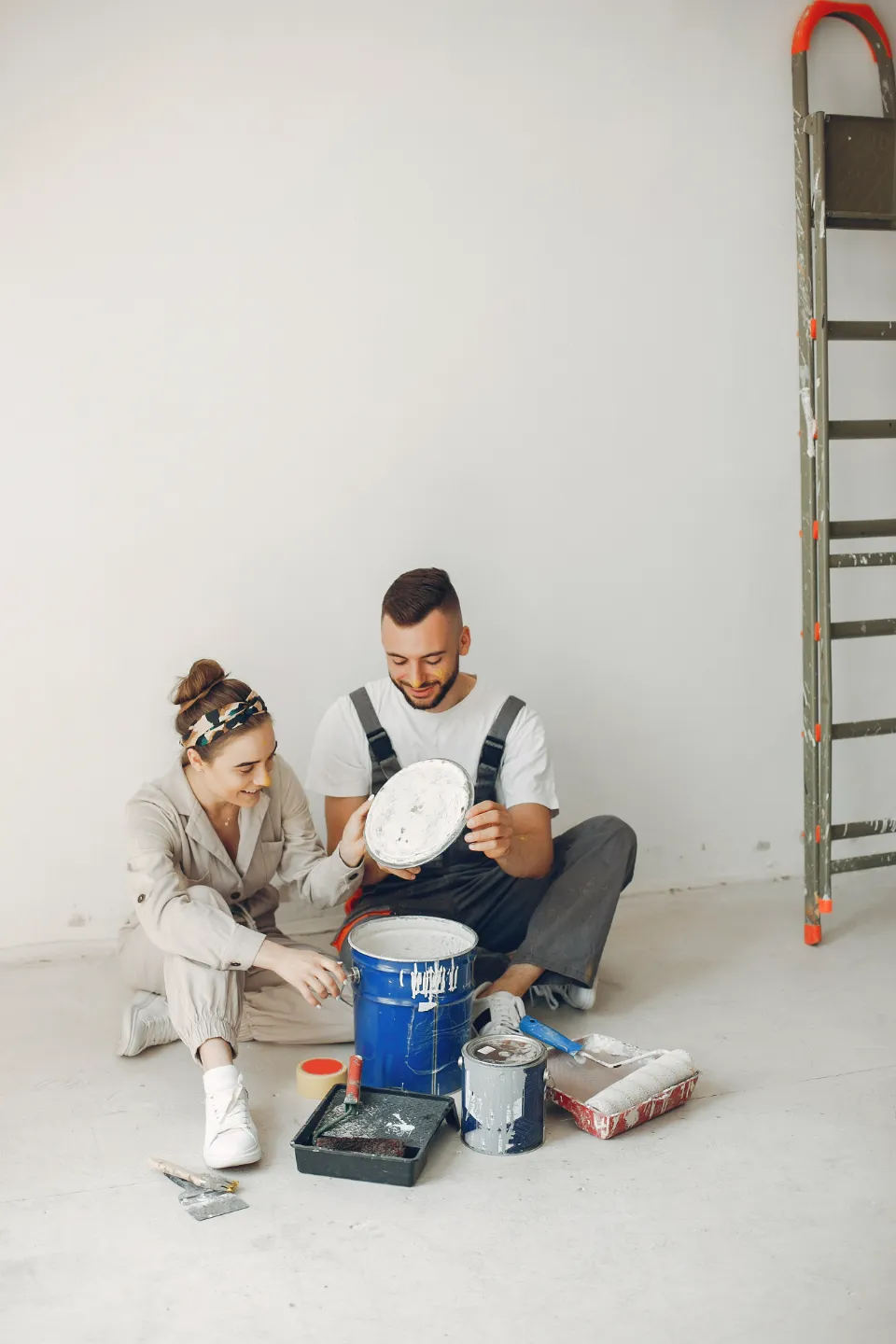 Un couple, habillé en salopette, s'apprêtant à peindre un mur. Ils sont assis au sol entourés de pots de peinture et d'outils.