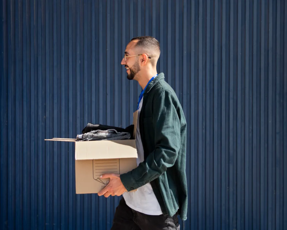 Homme souriant marchant sur le côté, portant une boîte en carton remplie d'objets, devant un mur bleu rayé.