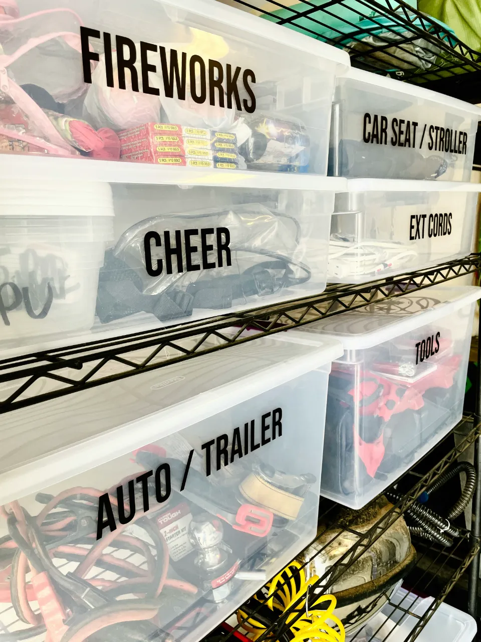 Plusieurs boîtes de rangement transparentes étiquetées, contenant divers articles comme des feux d'artifice, des affaires de cheerleading, des cordons d'extension, et du matériel pour voiture ou remorque.