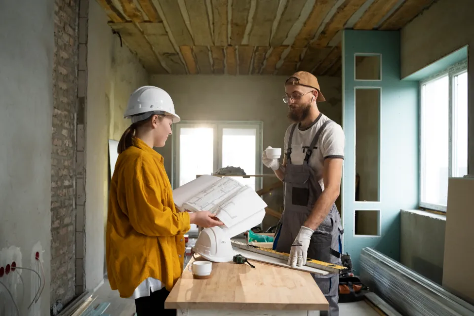 Deux ouvriers en casque, un homme et une femme, discutent des plans sur un chantier en rénovation.